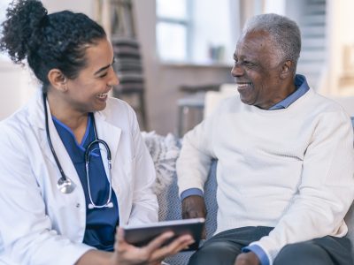 A female physician is meeting with a patient in the patient's home. The patient is a black senior adult man. The doctor and patient are seated next to each other on a couch. The doctor is holding a wireless tablet computer. The two individuals are smiling and looking at each other.